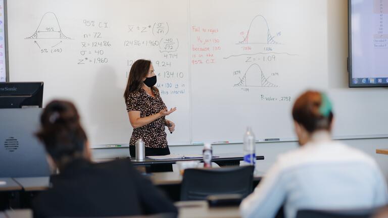 Math professor standing in front of a white board in a classroom.