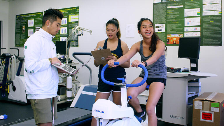 In a kinesiology lab a student rides a bike while two others take notes.