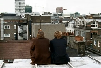 Screengrab from: A Refugee (2025) of two people sitting with their backs toward the camera on a roof overlooking the city on a cloudy day