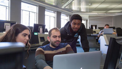 Three students huddled around a laptop in computer lab