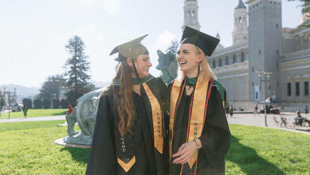 Two graduating honor students laughing in front of Wolf and Kettle sculpture