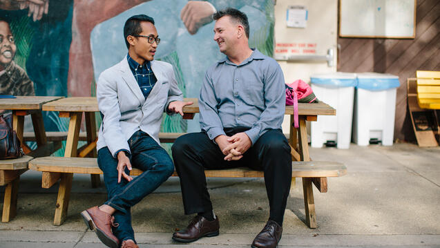 Two people sit on a bench in front of a mural and talk