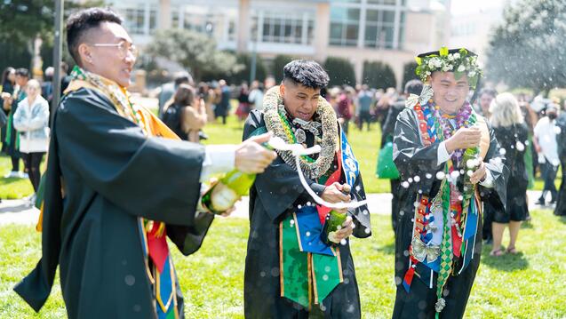 Three students wearing graduation regalia pop champagne on the lawn