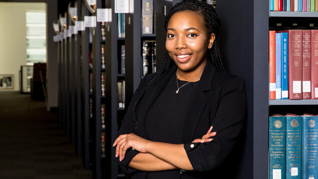 Student poses with crossed arms against a bookshelf.