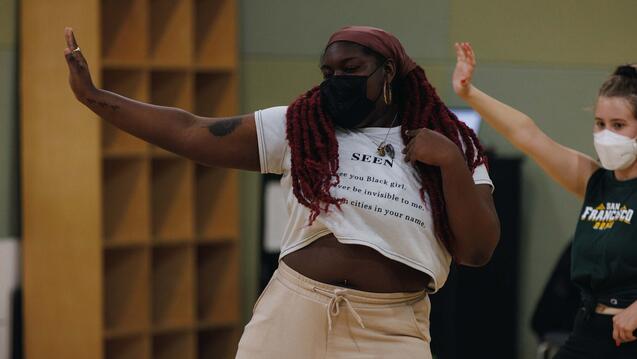 Dance student practices in studio with one arm pointing to the side and the other touching their shoulder