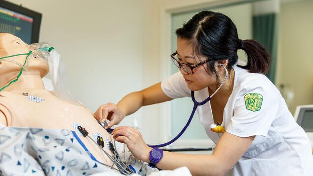 nursing student in scrubs holds stethoscope to manikin's chest