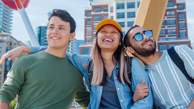Three students sit with arms around each other in front of bow and arrow sculpture downtown.