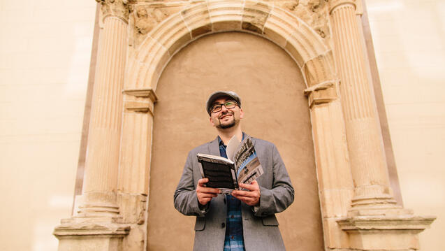 Professor holds out an open book while standing in front of an ornate archway.