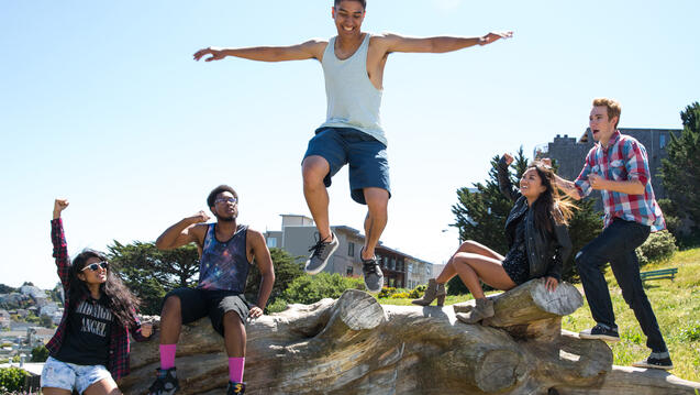 Four students sitting on a log cheer a person on who happily jumps off of the log