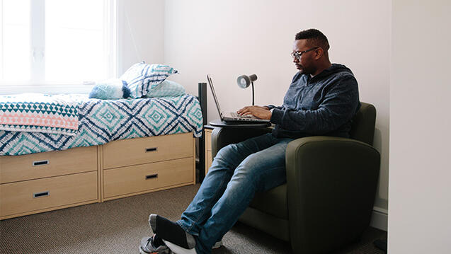 Grad student in dorm room working on laptop next to colorful bed