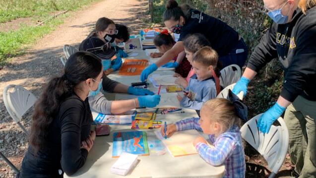 Students and teachers read books and complete art projects while sitting at a table outside.