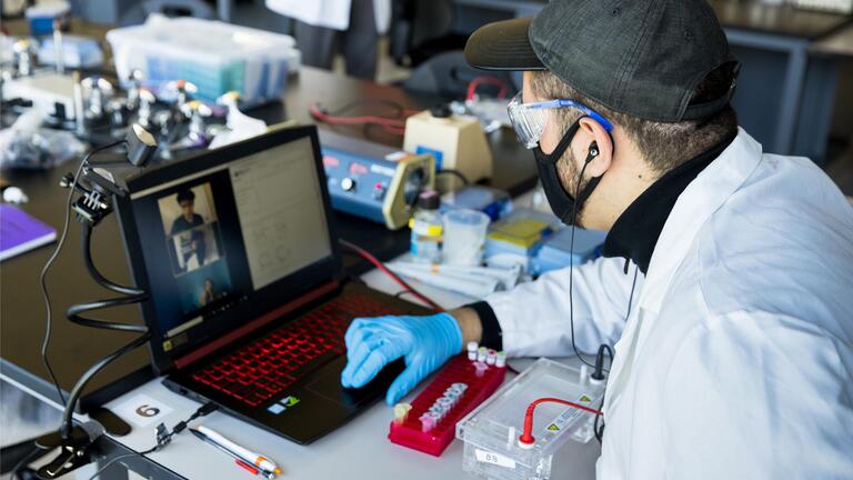 Lab associate in protective gear sits at desk with vial tray and joins video conference on laptop