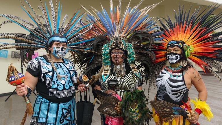 Three people celebrating Dia de Los Muertos in traditional attire with colorful feathered headdresses