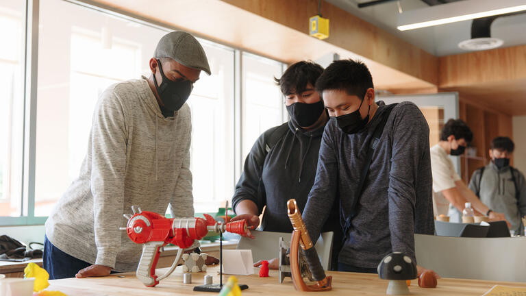 Three students look at a table with engineering experiments on it.