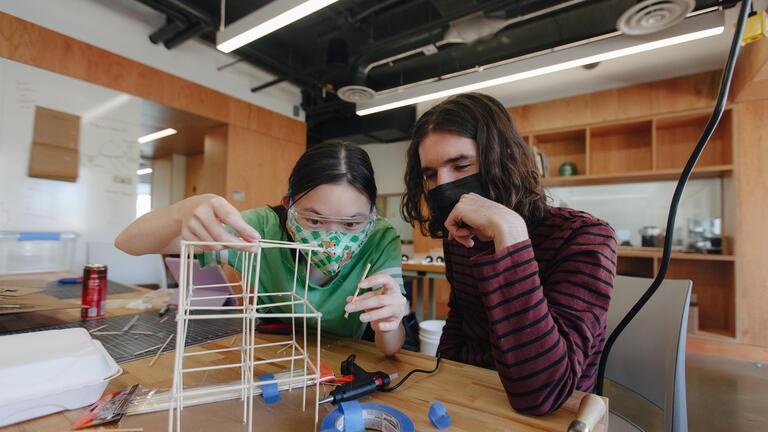 Two students build frame using wooden sticks, masking tape, and hot glue gun