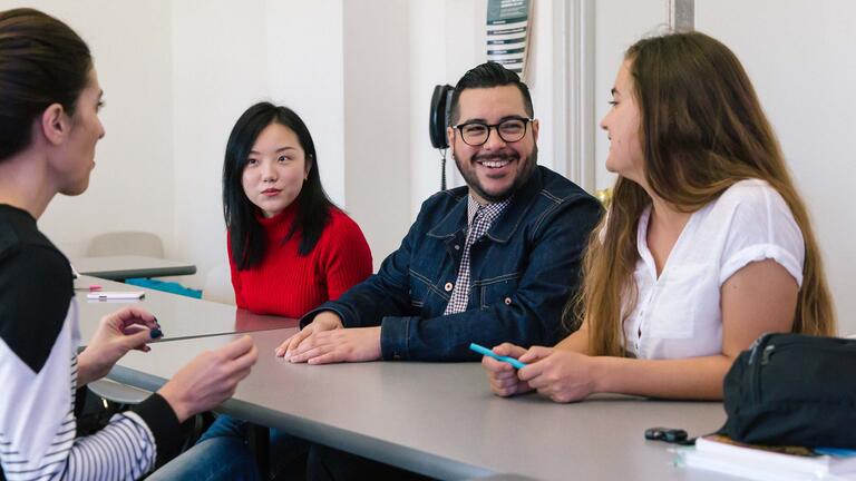 A group of students at a table speak to their professor.