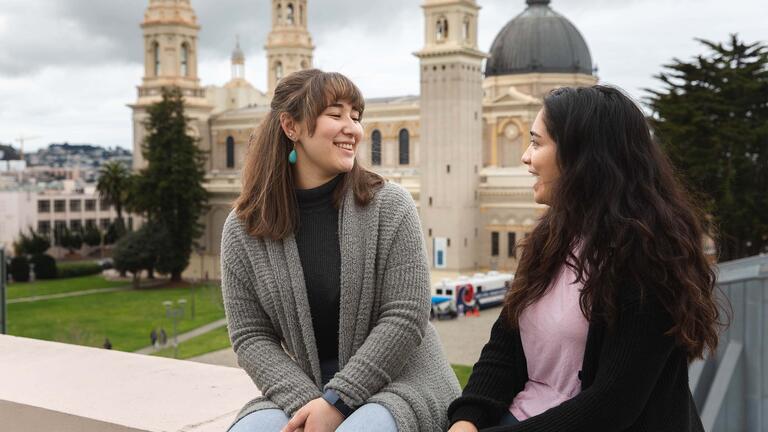 Two students chat while sitting on a wall.