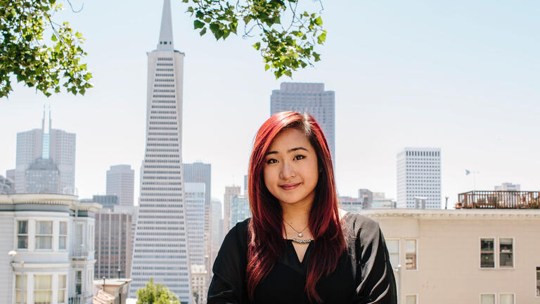 Student leans against a railing with the city skyline behind.