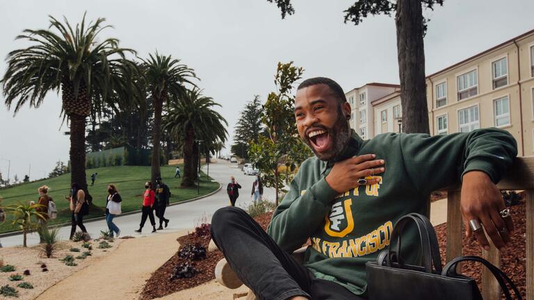 Student in USF sweatshirt sits on bench in front of Lone Mountain dorms.