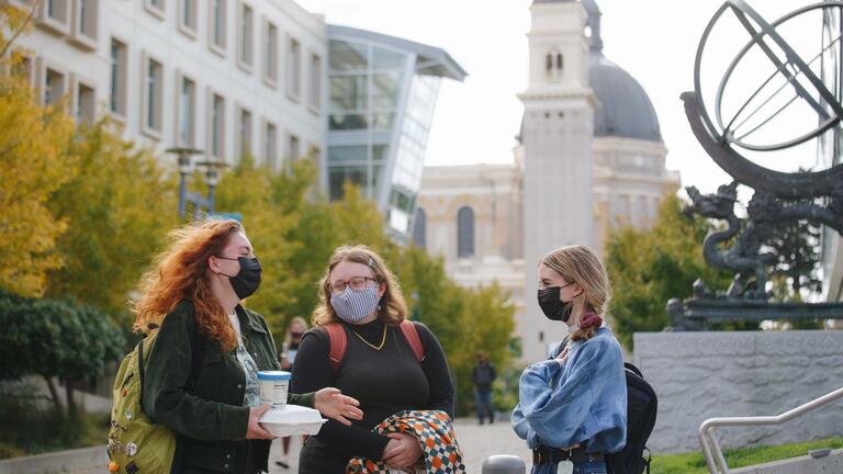 Three students talk on lower campus with the church in the background.