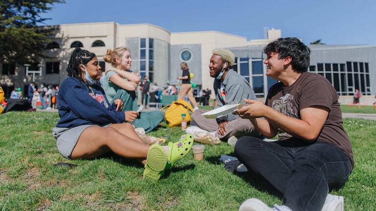 Students eat lunch and laugh on the lawn.