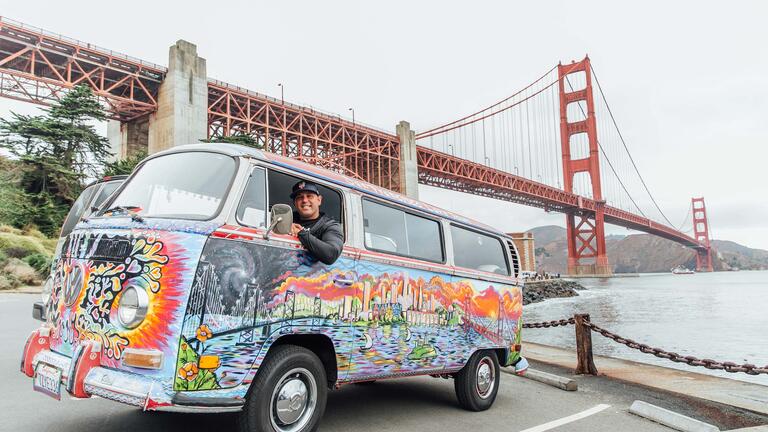 A colorfully painted mini van is parked under the Golden Gate Bridge.