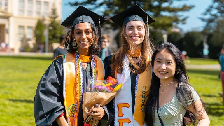 Two students wearing graduation regalia stand in the lawn and pose for a picture with a friend