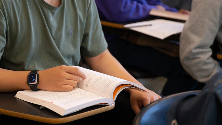 Torso and arms of student sitting at a desk with one hand on an open book