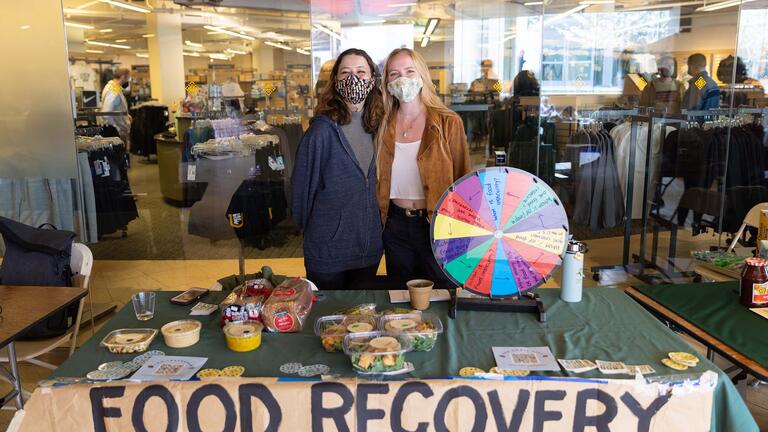 Two students stand next to each other behind a table with food items on it and a hand-drawn sign that reads “Food Recovery Network”
