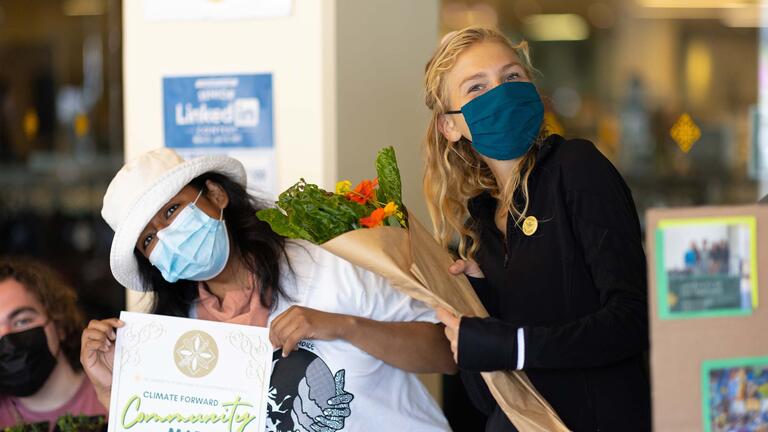 Two students pose for a picture, one holding flowers and the other a sign that reads “Climate Forward Community Market”