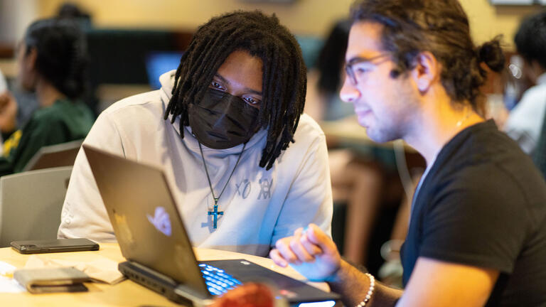 Two students sit at a table and collaborate in front of an open laptop compute