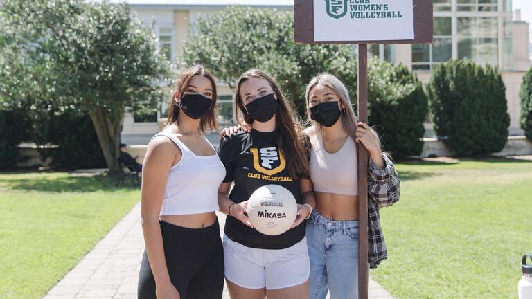 Three students on Welch Field hold a sign for USF Club Women's Volleyball.