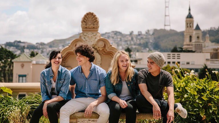 Four students sit on a bench with a view of campus in the background.