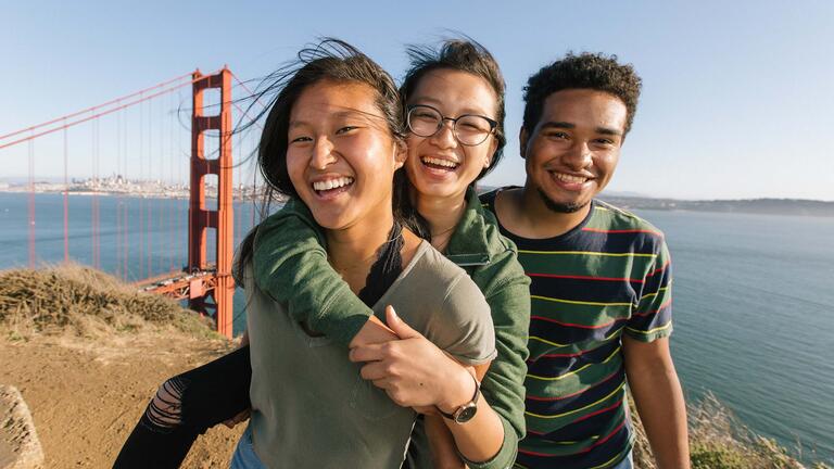 Three students embrace and smile on a hilltop overlooking the Golden Gate Bridge.