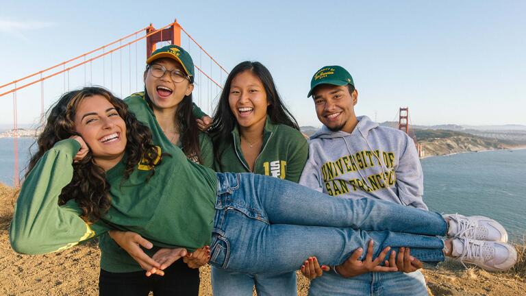 Three students hold another student in their arms on a hill with the Golden Gate Bridge behind.