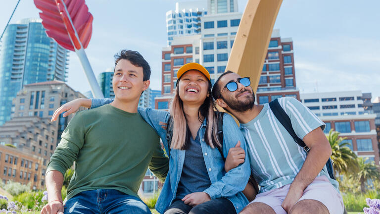 Three students embrace in front of a bow and arrow sculpture in San Francisco.