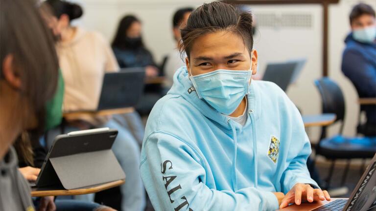 Student sitting at a desk leans sideways to talk to a classmate