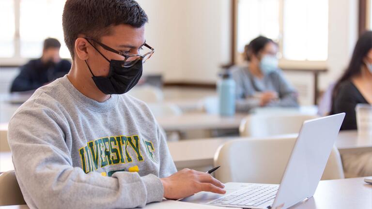 Student sits at a desk in class working on a laptop with an open notebook next to it