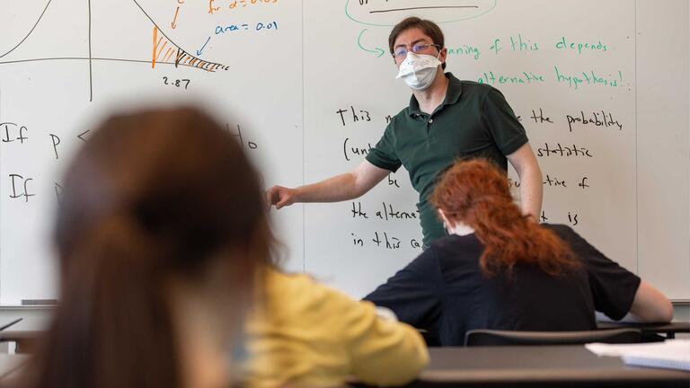 Statistics professor reaching with right arm explains the graph on the white board behind