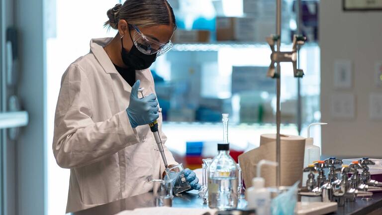 Student wearing protective gear holds a syringe in a beaker with measurements in the lab