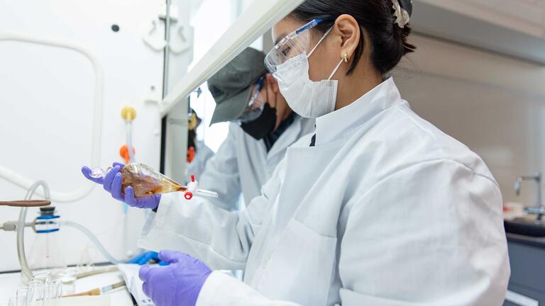 Two students in protective gear work with beakers inside the biosafety cabinet