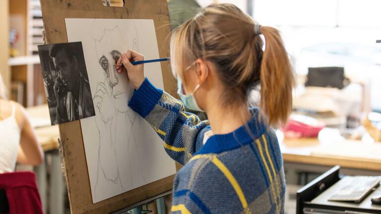 Student at an easel works on a pencil drawing while using a photograph as a reference