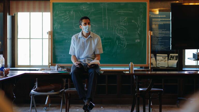 Instructor sits on a desk lecturing in front of a chalkboard