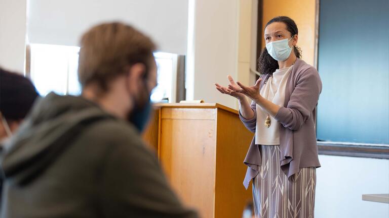 Instructor lectures in front of a podium in class while gesturing with hands