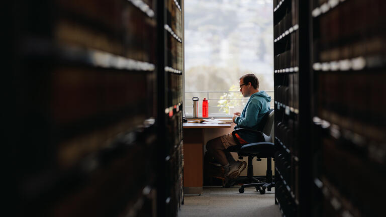 Student seen through a row of bookshelves studies at a table.