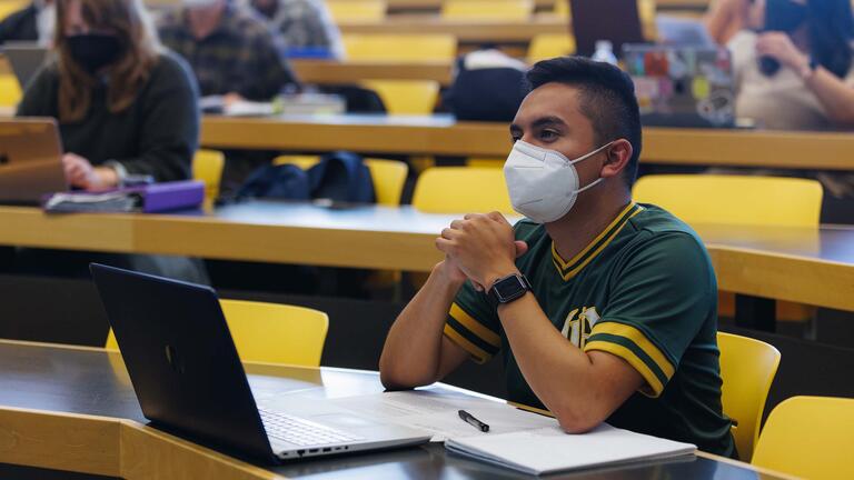 Student with a laptop listens with elbows on the table and hands clasped in the lecture hall