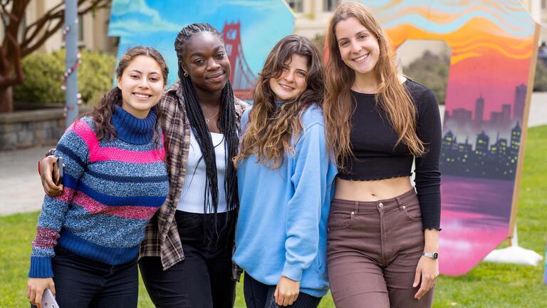 Four students pose in front of a colorful mural that reads DONS.