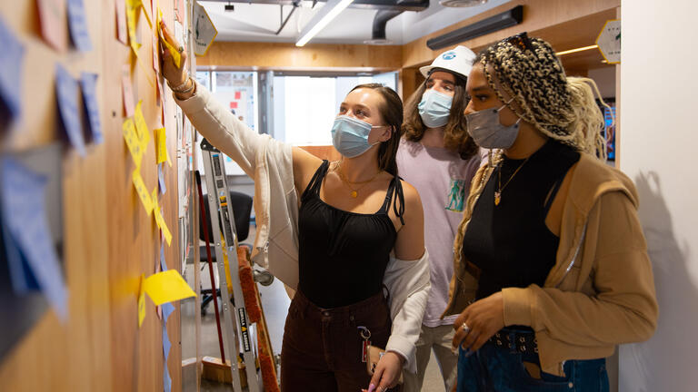 A student shows other students research documentation pasted on a wall.