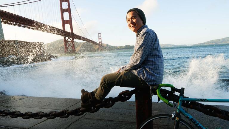 Student sits on a post at the edge of the bay with the Golden Gate Bridge behind him.