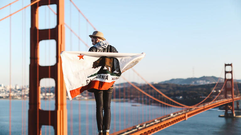 Student wrapped in a California flag stands on a high point with the Golden Gate Bridge behind.